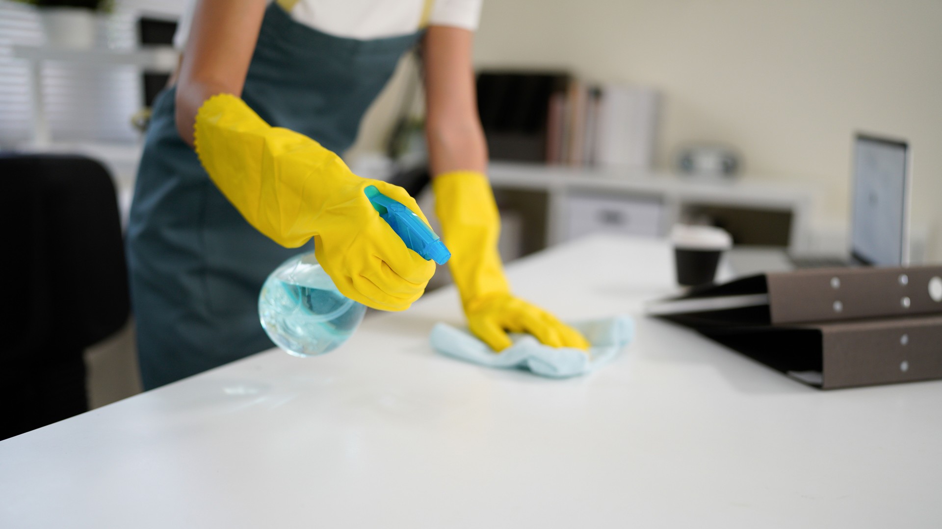 Young asian woman wearing yellow gloves and an apron, smiling while cleaning a white table with disinfectant spray and a blue cloth, promoting hygiene and cleanliness in her workspace